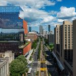 Photo: View of Boston University's urban campus featuring a striking multi-layered glass and metal building, tree-lined streets with tram tracks, and high-rise buildings in the background under a partly cloudy sky.