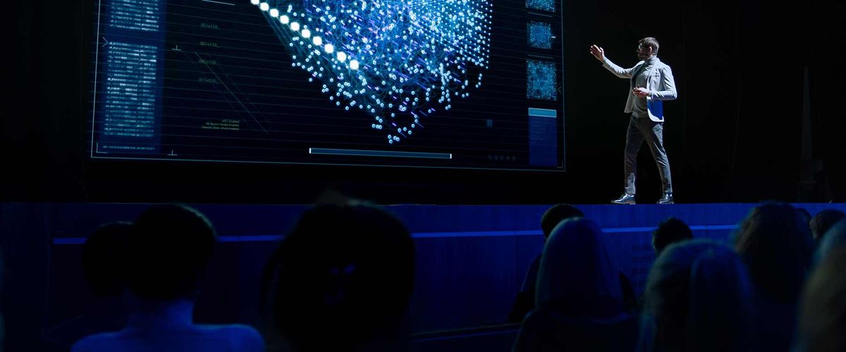 A speaker on stage at a technology conference, presenting graphics on a large screen