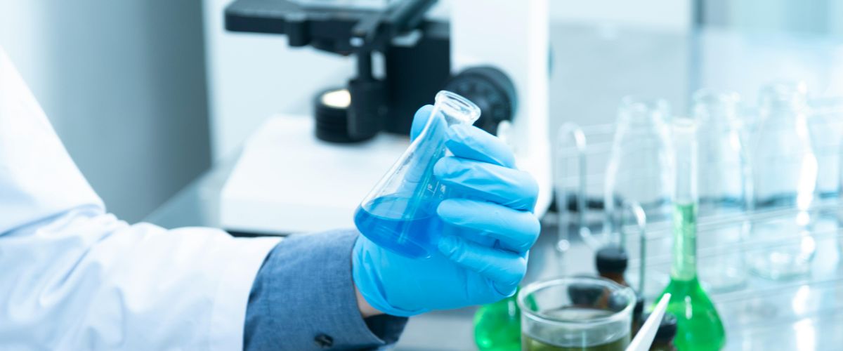 Person in lab coat and gloves holding a beaker with blue liquid in it