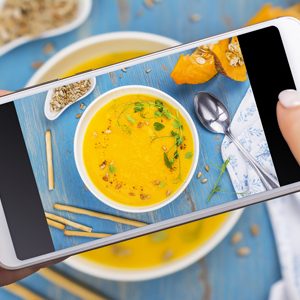 Woman taking photo of fall spicy pumpkin soup with herbs, croutons and sunflower seeds. Trendy instagram photo shot. Top view, flat lay style. Copy space