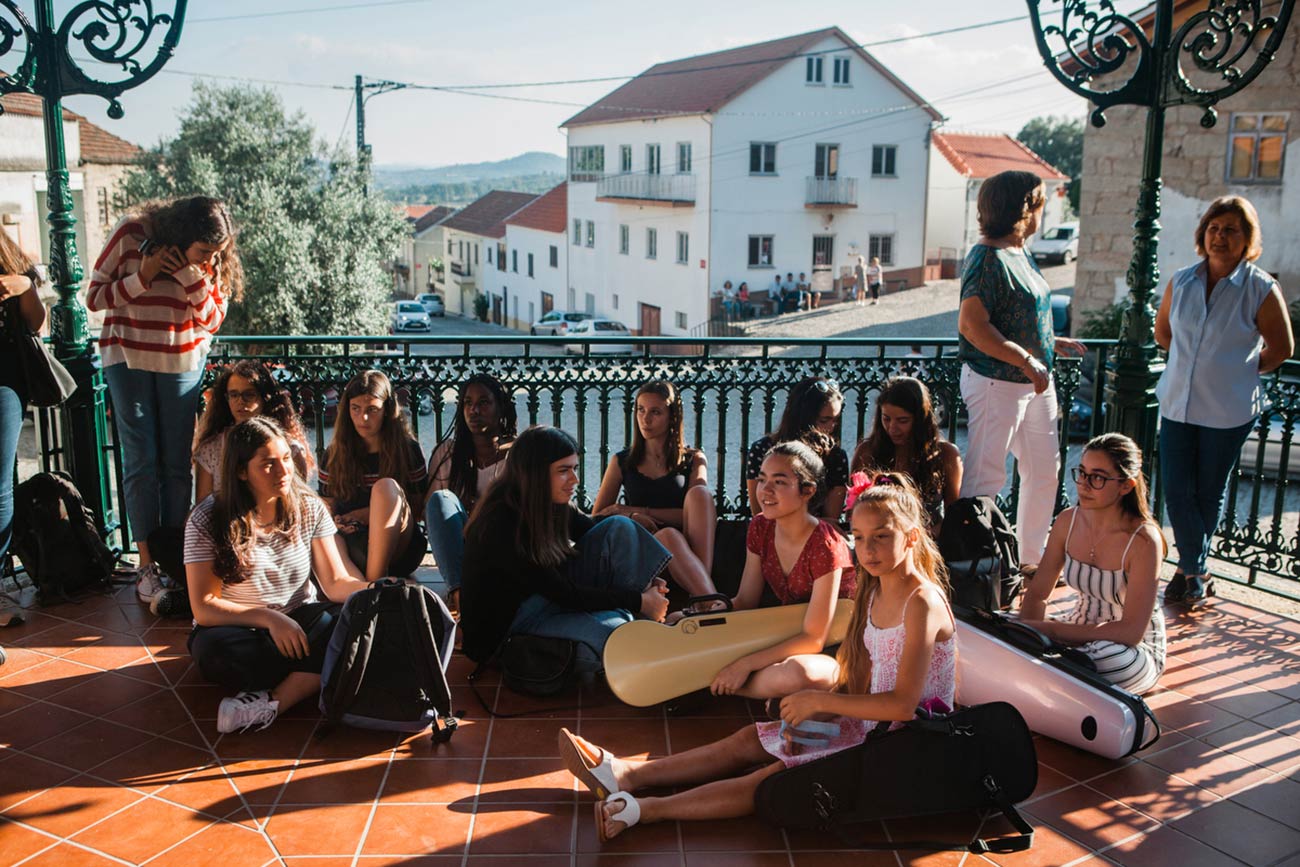 Full color slideshow photo of Bendada Music Festival students and Bendada villagers gathered sitting and standing in the shade of the coreto bandstand watching a festival performance.