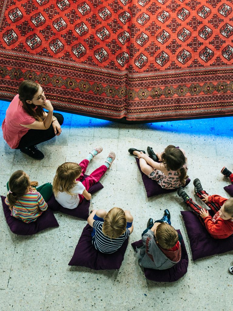 A woman crouching, surrounded by children sitting on pillows. There is a Persian style rug laid out in front of them.