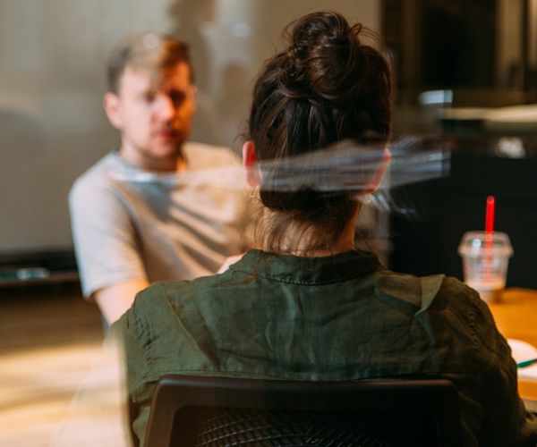 A male presenting person and a person with a bun whose back is to us sit behind glass and talk.