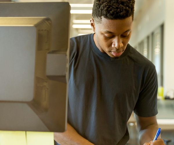 A young black man stands at a computer and writes on paper