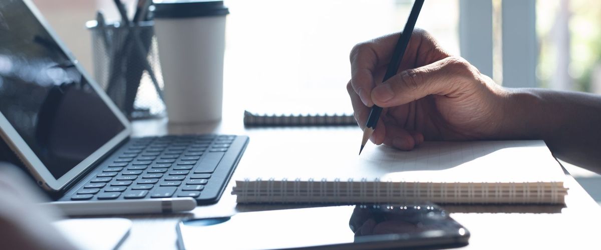 Photo of a person's hand taking notes in front of a laptop