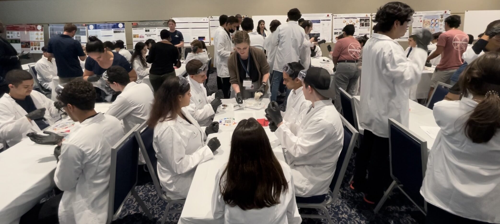 A CELL-MET trainee is standing up holding out a pig's heart as she uses her finger to point to where a valve is. Five high school students in lab coats are around her watching intently.