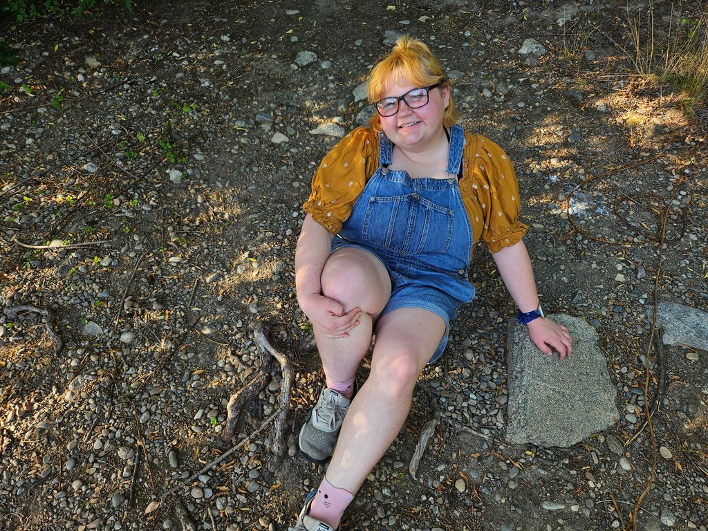 A person with blonde hair, glasses, and a warm smile, sitting outdoors on a rocky surface. They are wearing denim overalls, a mustard yellow shirt with puffed sleeves, and sneakers, appearing relaxed and joyful in a natural setting.