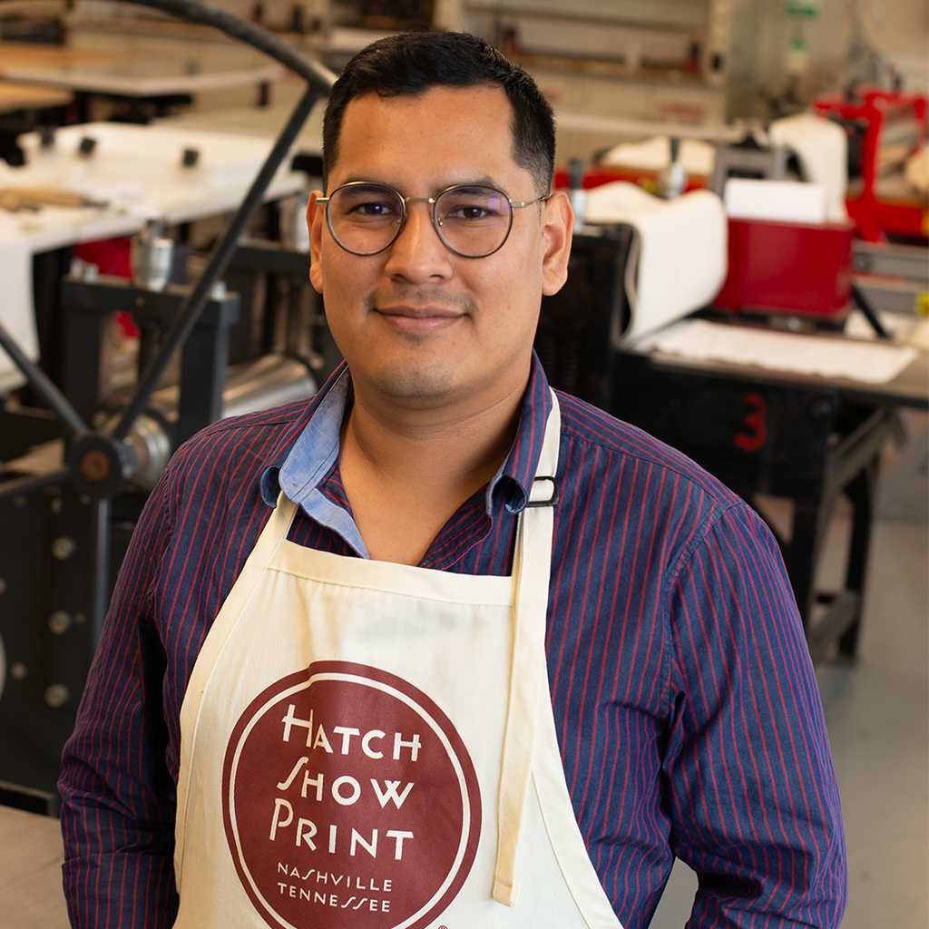 Jerry Rodriguez Sosa, a BU MFA Print Media & Photography student, is pictured in BU School of Visual Arts' Printmaking Studio. Jerry is smiling at the camera, sporting a long sleeved purple striped collar shirt, round glasses, and wearing a beige apron that says, "Hatch Show Print. Nashville, Tennessee."