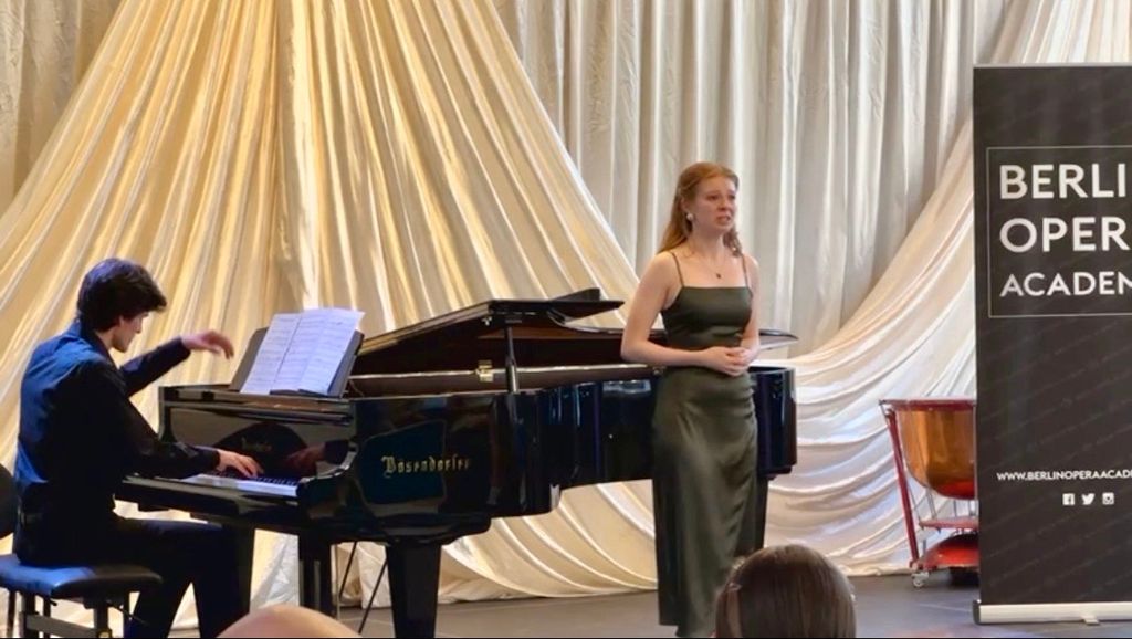 A young woman in a green dress performs a vocal piece beside a pianist seated at a black Bösendorfer grand piano. Behind them, a backdrop of elegant draped curtains and a sign reading 'Berlin Opera Academy' is visible.
