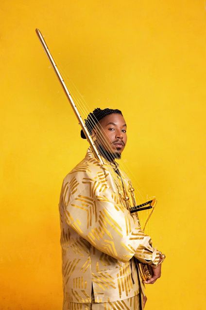A person wearing a gold and cream patterned outfit poses against a bright yellow background, holding a stringed musical instrument with golden accents. The individual has braided hair styled neatly and looks confidently toward the camera, creating a vibrant and artistic composition.