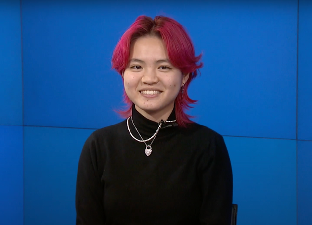 BU College of Fine Arts BA music major Miya Menscher with vibrant pink, layered hair sits in front of a bright blue background, smiling warmly at the camera. They are wearing a black turtleneck sweater, accessorized with layered silver necklaces, one featuring a heart-shaped pendant. They also wear dangling earrings, adding to their stylish look.