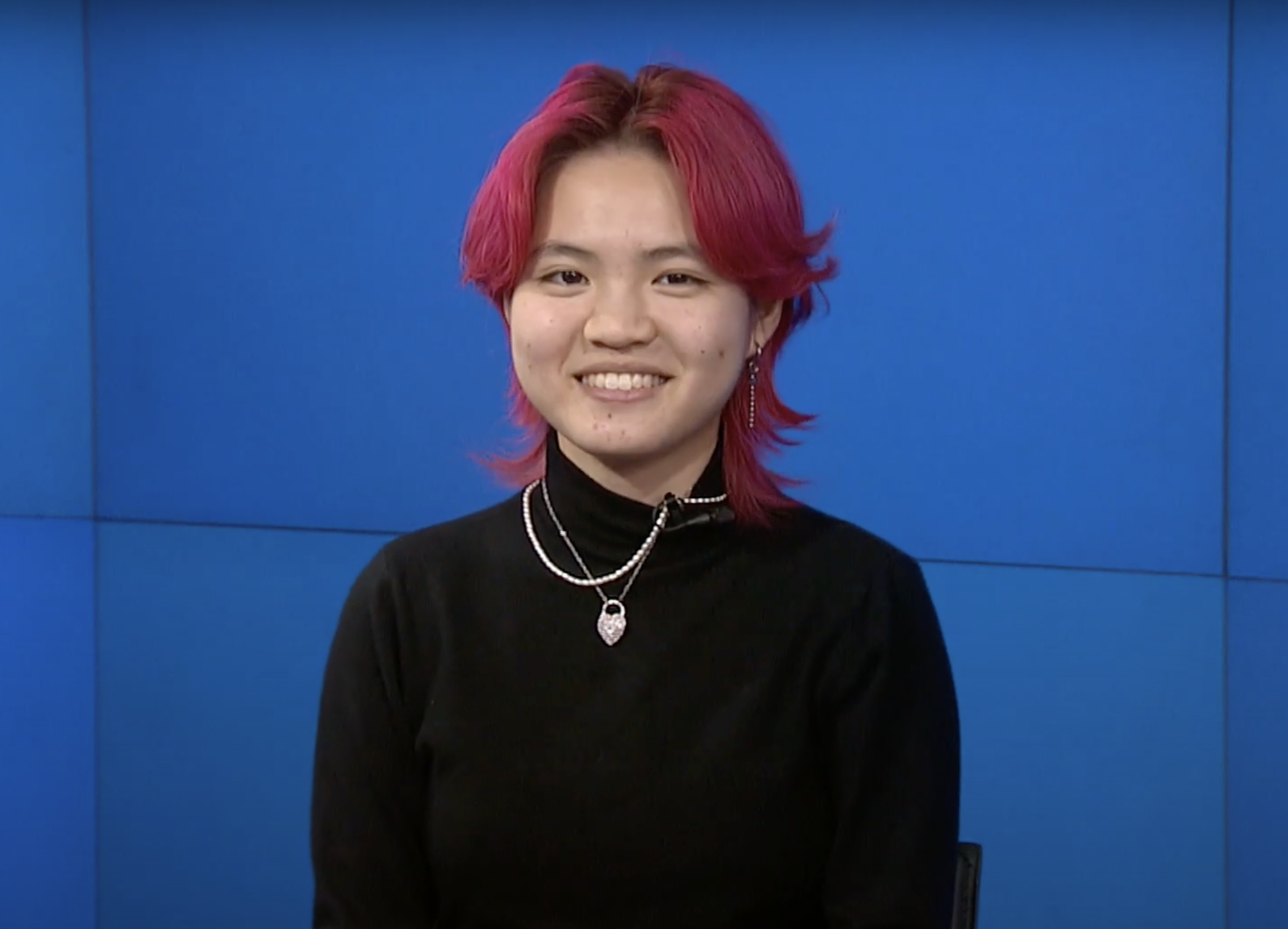 BU College of Fine Arts BA music major Miya Menscher with vibrant pink, layered hair sits in front of a bright blue background, smiling warmly at the camera. They are wearing a black turtleneck sweater, accessorized with layered silver necklaces, one featuring a heart-shaped pendant. They also wear dangling earrings, adding to their stylish look.