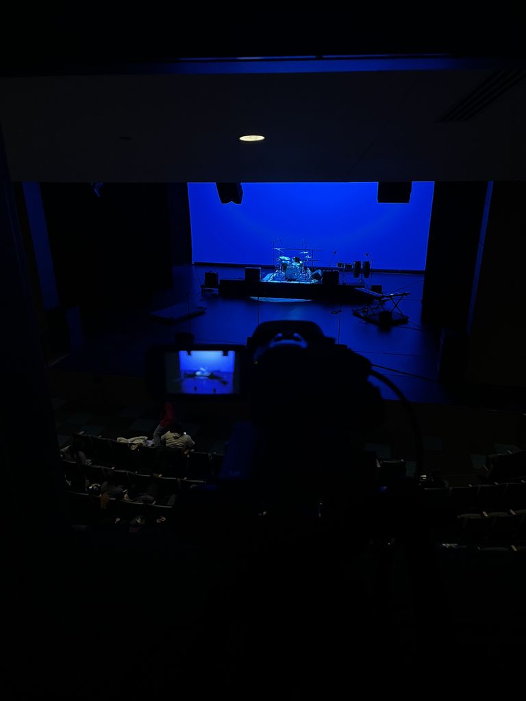 A dimly lit theater view from the back of the audience, looking down at a stage illuminated by a deep blue backdrop. A drum set, microphones, and other musical instruments are set up on stage, ready for a performance. A camera in the foreground is capturing the scene, with its small screen displaying the same stage view. A few audience members are seated, waiting for the event to begin.