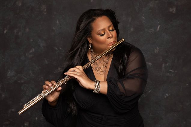 Grammy nominated composer Valerie Coleman, with long, flowing black hair, plays a golden flute against a dark, textured background. She is wearing a black sheer-sleeved dress, gold jewelry, and has shimmering gold eyeshadow. Her expression is focused and passionate as she performs, with her fingers elegantly positioned on the flute's keys.
