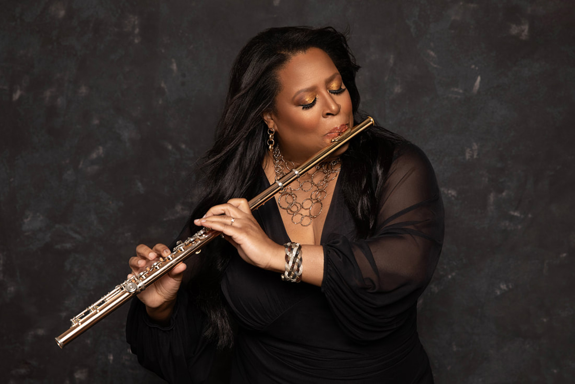 Grammy nominated composer Valerie Coleman, with long, flowing black hair, plays a golden flute against a dark, textured background. She is wearing a black sheer-sleeved dress, gold jewelry, and has shimmering gold eyeshadow. Her expression is focused and passionate as she performs, with her fingers elegantly positioned on the flute's keys.