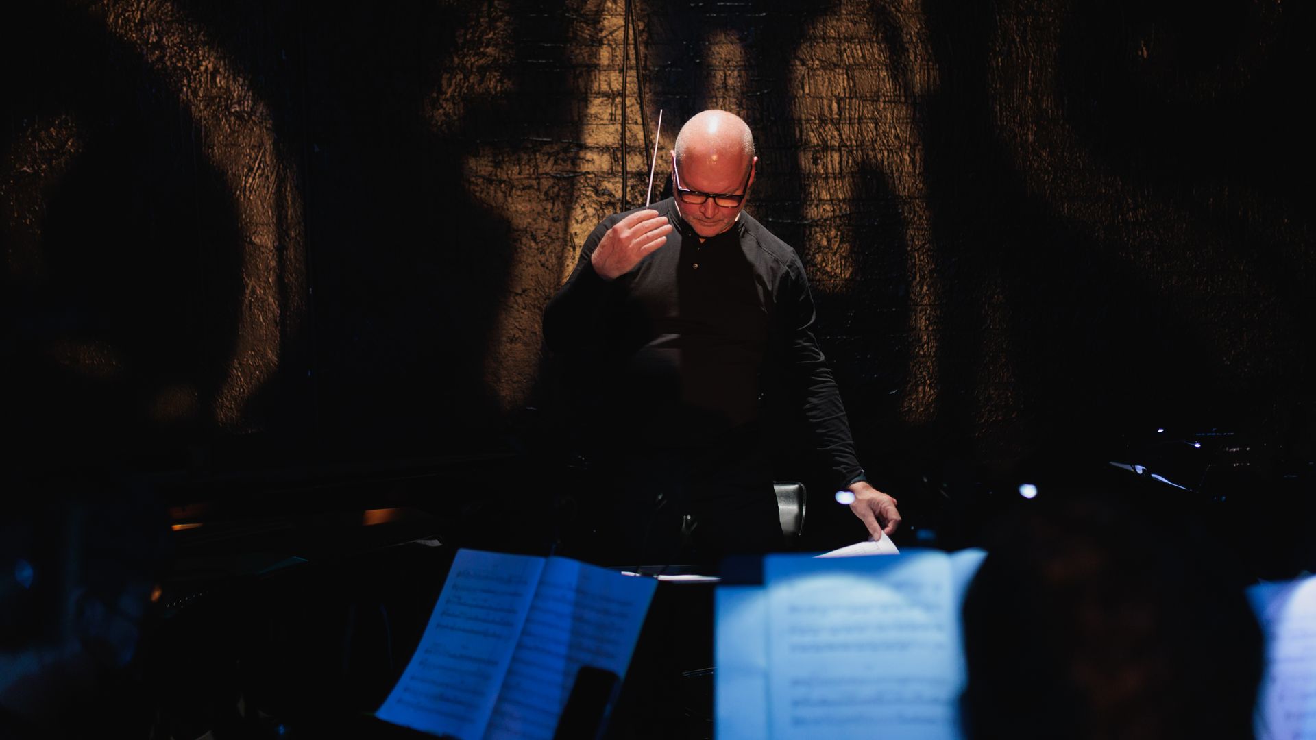 BU Opera Institute Artistic Director William Lumpkin dressed in black leads a musical performance in a dimly lit setting. He stands in front of an orchestra, raising one hand while holding a baton, with an intense and focused expression. The foreground features blurred sheet music illuminated by soft blue light. The background consists of a textured, dark wall with dramatic shadows, creating a moody and atmospheric scene