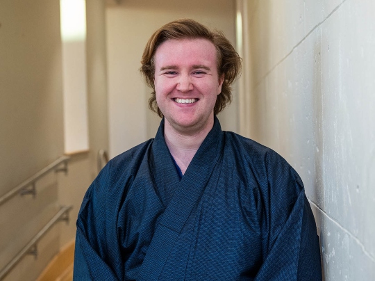 Christopher Ellars (CAS’24, CFA’25), a dual Japanese and voice performance major with medium-length, wavy blond hair stands in a softly lit hallway, smiling warmly at the camera. He is wearing a traditional dark blue Japanese kimono with a red obi belt. The background features cream-colored walls, a wooden floor, and handrails along the sides, giving the space a serene and elegant atmosphere.