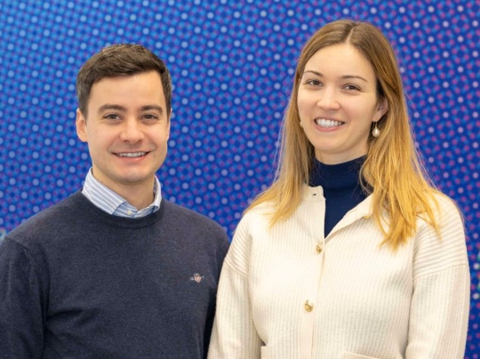 Edoardo Carpenedo (CFA’19) (left) and Inês Andrade (CFA’18) standing side by side, smiling at the camera. Edoardo is wearing a navy blue sweater over a collared shirt, while Inês is wearing a cream-colored cardigan over a navy blue turtleneck. They are posed against a vibrant blue background with a dotted pattern, giving the image a lively and colorful atmosphere.