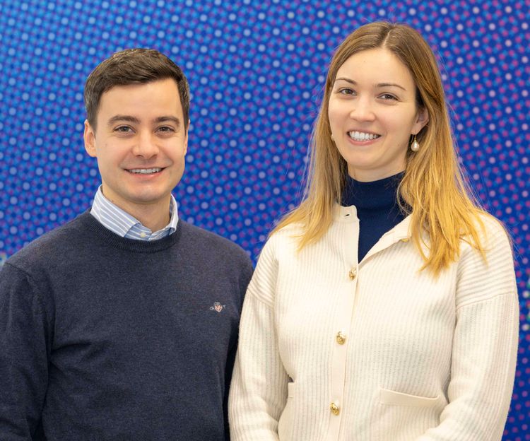 Edoardo Carpenedo (CFA’19) (left) and Inês Andrade (CFA’18) standing side by side, smiling at the camera. Edoardo is wearing a navy blue sweater over a collared shirt, while Inês is wearing a cream-colored cardigan over a navy blue turtleneck. They are posed against a vibrant blue background with a dotted pattern, giving the image a lively and colorful atmosphere.