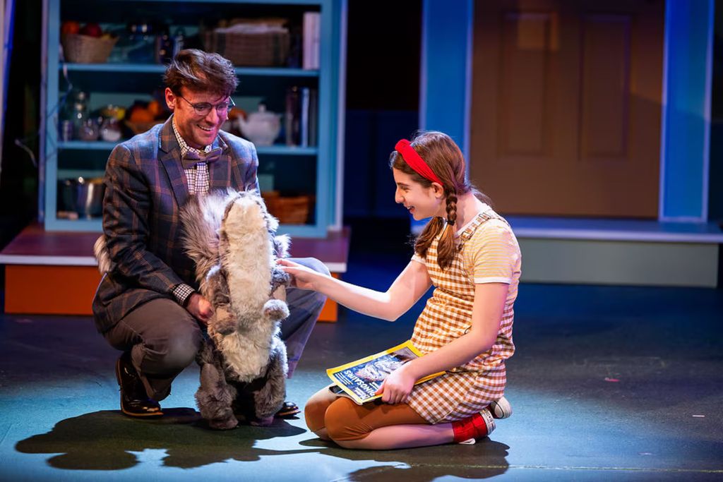 A scene from Wheelock Family Theatre’s production of Flora & Ulysses featuring two characters interacting with a large squirrel puppet. On the left, a man dressed in a plaid blazer and bow tie kneels, smiling warmly as he holds the puppet. On the right, a young girl in a yellow checkered dress, red headband, and braided hair kneels, joyfully petting the squirrel puppet while holding a book. The background is a vibrant, colorful set designed to resemble a cozy living room. The scene captures a playful and heartwarming moment, highlighting the whimsical nature of the performance.