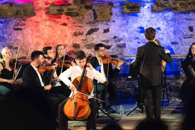 A cellist performs passionately at the center of an orchestral ensemble, surrounded by violinists and other string musicians. The conductor, dressed in a black suit, leads the orchestra with expressive gestures. The backdrop features a rustic stone wall illuminated with vibrant red and blue lighting, creating a dramatic and dynamic atmosphere that enhances the performance's emotional impact.