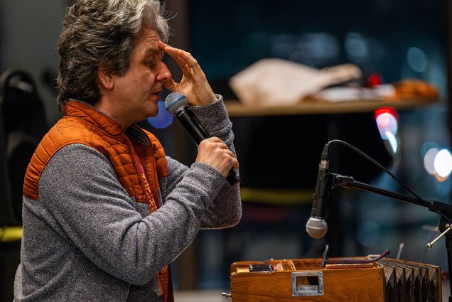 CFA’s Igor Iwanek seen with wavy gray hair and light skin holds a microphone while speaking at one of the breath workshops he leads, with his other hand resting on his forehead. He is wearing a gray sweater with an orange quilted vest. In front of him is a wooden harmonium with a microphone positioned nearby. The background is blurred, with warm indoor lighting and hints of a reflective surface, possibly a window or glass wall, with city lights visible outside.