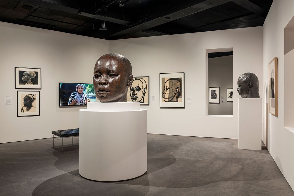 Witnessing Humanity: The Art of John Wilson exhibition at the Museum of Fine Arts, Boston. The exhibition showcases a prominent bronze sculpture of a head titled "Eternal Presence" on a large white pedestal at the center. Surrounding the sculpture are framed sketches and portraits depicting various perspectives of the head, highlighting the artistic process and exploration of form. A smaller bronze head is displayed on a pedestal in an adjacent room, visible through a wall opening. A video screen shows a documentary interview, adding contextual depth to the exhibition. The room is softly lit, emphasizing the detailed textures and expressive qualities of the artworks.