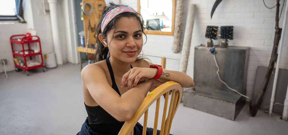 Maithili Rajput, a Boston University graduate sculpture student, with dark hair tied back with a colorful headband sits backward on a wooden chair in an artist’s studio. She wears a black tank top, navy work pants, a red watch, and wireless earbuds, smiling gently at the camera. The background features a white brick wall adorned with sculptural tools and materials, including a large black metal scythe, wooden logs with spikes, and a workstation with various equipment. Sunlight streams in from a window, casting a warm glow on the creative workspace.
