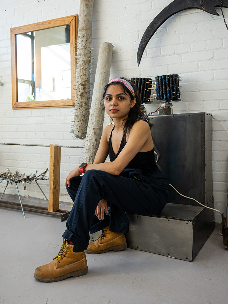 Maithili Rajput, a Boston University graduate sculpture student, with dark hair, wearing a black tank top, navy work pants, and tan work boots, sits on a metal sculpture in an industrial-style studio. She has a pink and white headband and a red smartwatch on her wrist. The studio has a white brick wall with various sculptural pieces, including spiked wooden poles, a large curved blade, and an electronic component mounted on the wall. A wooden-framed mirror reflects a window, letting in natural light. She gazes at the camera with a calm and confident expression.
