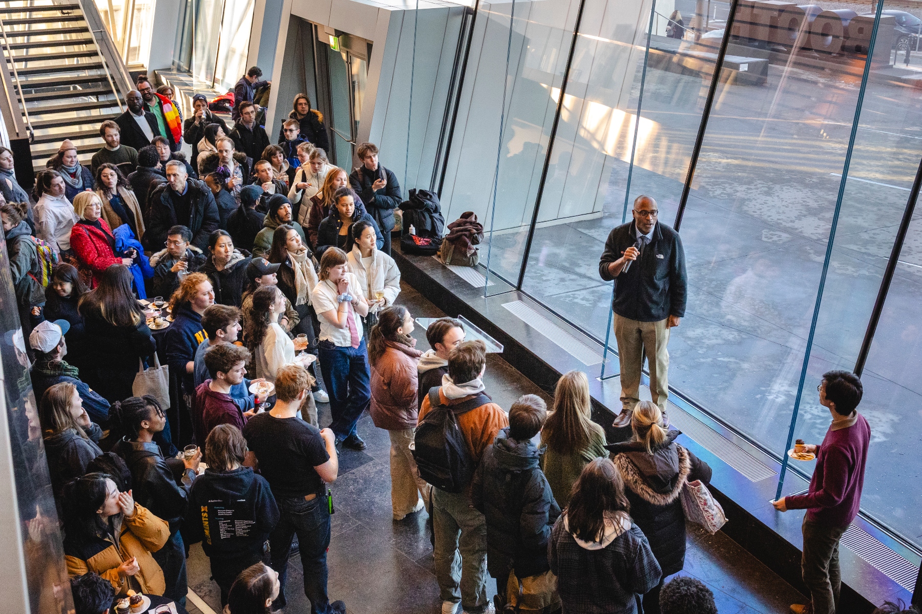 A large group of people gathers inside the Boston University Joan & Edgar Booth Theatre Yan Luo Lobby, a modern, glass-walled atrium, attentively listening to BU College of Fine Arts Dean Harvey Young. Dean Young is wearing glasses and a dark jacket, and stands near the floor-to-ceiling windows holding a microphone. The audience, consisting of CFA students, faculty, and staff of various ages and diverse backgrounds, is dressed in warm clothing, with some holding plates of food and drinks. The natural light from the windows casts a glow on the space, and reflections can be seen on the glass.