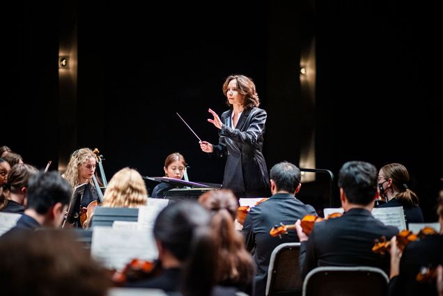 Conductor Sarah Ioannides leads the Boston University Symphony Orchestra in performance. She stands in front of the ensemble wearing a black suit, baton in hand, mid-gesture. The musicians, seen from behind and in profile, are intently following her direction in a dimly lit concert hall.