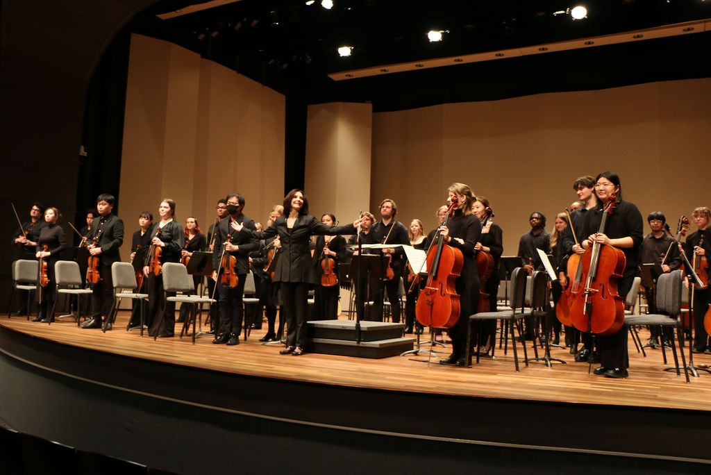 Conductor Sarah Ioannides stands center stage with a smile and an outstretched arm as the Boston University Chamber Orchestra rises for a bow at the end of a performance. Musicians dressed in black hold their string instruments, acknowledging the audience in a warmly lit concert hall.