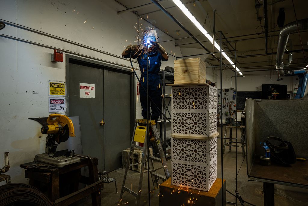 Maithili Rajput, a Boston University graduate sculpture student, wearing a protective helmet and gloves stands on a ladder in a workshop, welding a metal structure. Bright sparks fly from the welding point, illuminating the dark industrial workspace. In the foreground, a stack of intricately patterned white metal boxes with a wooden crate on top is visible. Various tools, machinery, and safety signs, including warnings for eye protection and heavy machinery, are present on the walls. The scene captures the energy and craftsmanship of metalworking in an industrial setting.