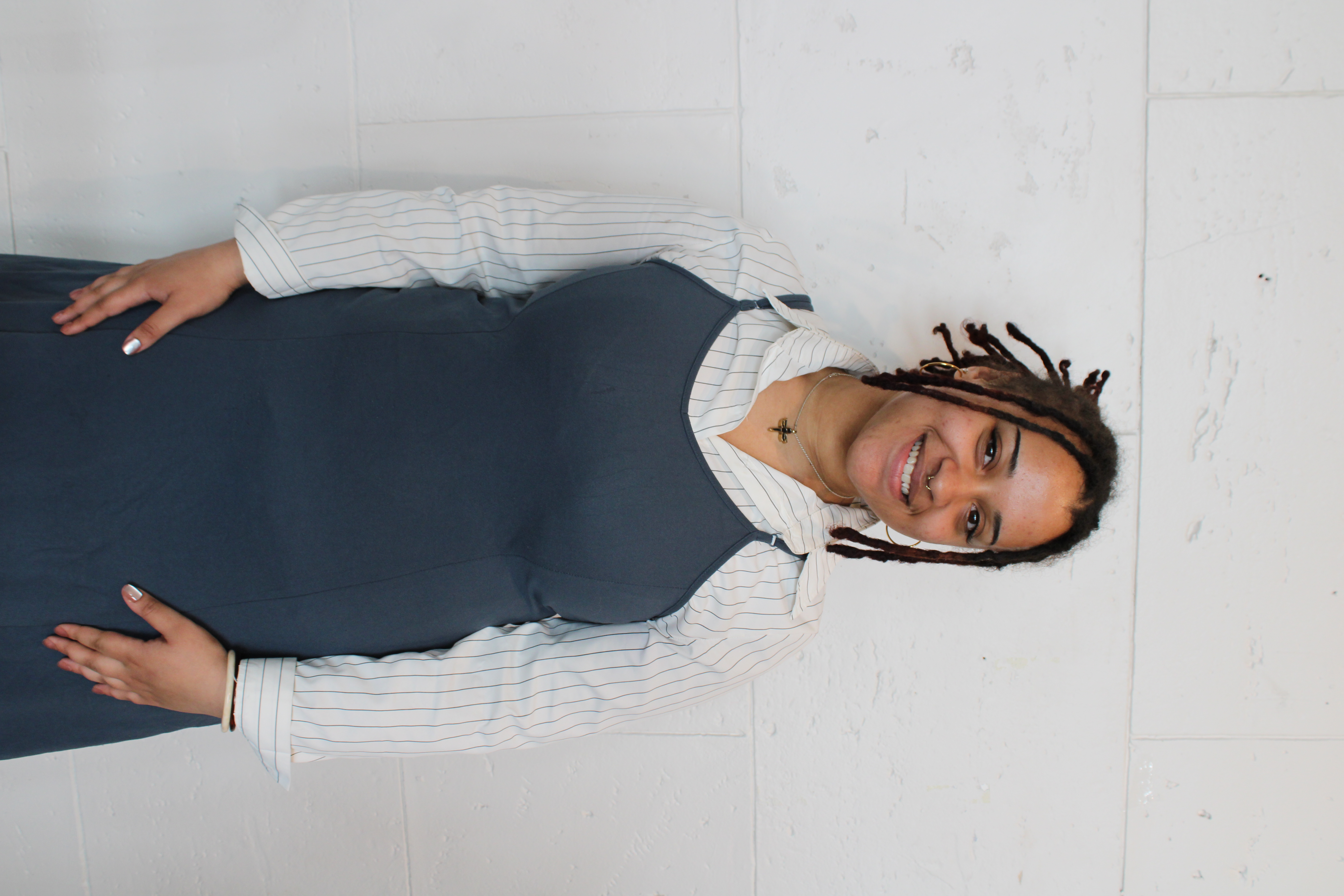 A portrait of Nyla Mawire standing against a white wall, smiling warmly at the camera. Nyla is a tuba performance undergraduate student at 波士顿大学 音乐学院 and the University's 2025 Kahn Career Entry Award recipient. She wears a blue dress layered over a white pinstriped shirt and has her hands loosely clasped in front of her.