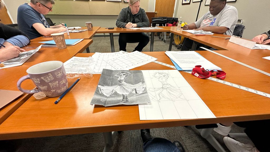 A group of adults seated around a rectangular table, working on sketches. The table holds sketching materials, photo references of a dancer, and grid templates. A lavender mug with butterfly prints, a BU lanyard, and rulers are also visible on the table.
