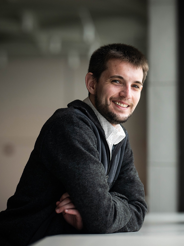 Portrait of 雅各布·惠特, a 波士顿大学 Master of Music in Composition student, smiling and leaning on a table with arms crossed, dressed in a dark sweater over a collared shirt, with soft indoor lighting and a blurred background.