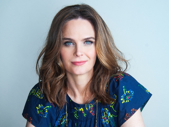 Actor Emily Deschanel with wavy brown hair and blue eyes sits at a table with her arms crossed, wearing a navy blue top with a colorful floral pattern and a long necklace, looking confidently at the camera against a light gray background.