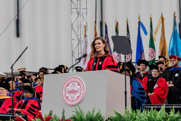 Actor Emily Deschanel speaks at a podium bearing the 波士顿大学 seal during the 2025 Commencement ceremony. She wears academic regalia as faculty members in caps and gowns sit behind her, with colorful flags displayed in the background.