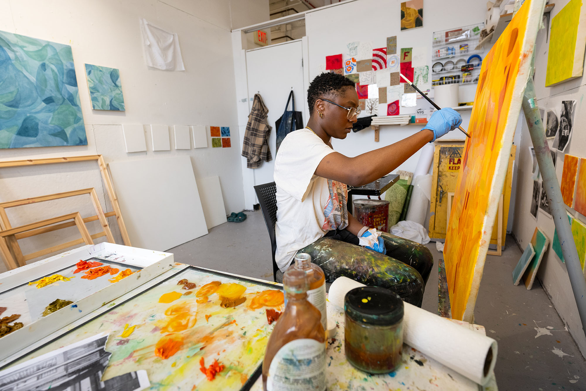 Raquel Philippe, a 波士顿大学 MFA Painting student, sits in her studio working on a large canvas in vibrant shades of orange. She wears paint-splattered pants, a casual graphic t-shirt, and blue gloves, focusing intently on her brushwork. Her studio is filled with colorful artwork, paint supplies, and blank canvases leaning against the wall.