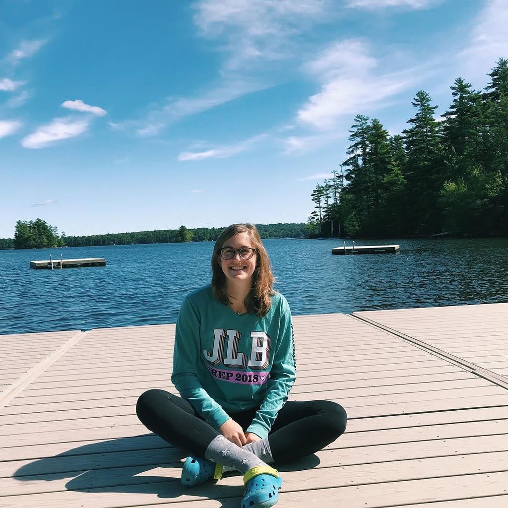 Caroline sitting on a pier with a lake, trees and a blue sky behind her