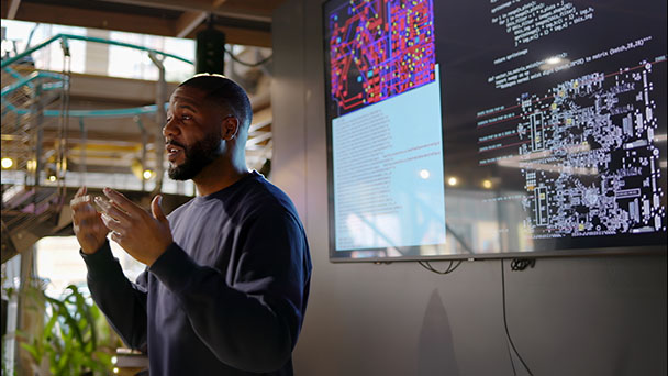 Black man speaking in front of a large screen.
