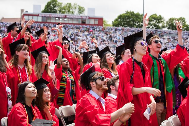 Group of graduates in BU regalia on Nickerson Field cheering, with a full bandstand of guests in the background.