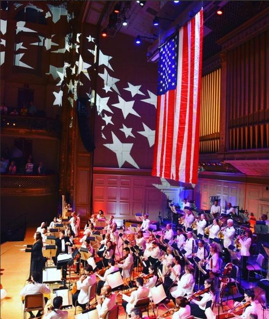 Boston Pops Orchestra performs under a large USA flag with star lighting on the walls