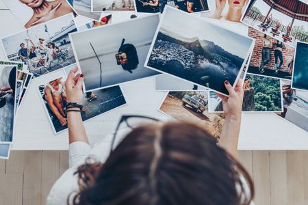 Overhead view of photographs spread out on a table. On the lower edge, a person can be seen: we look down at their brown hair and see their hands holding up two photographs from the pile.