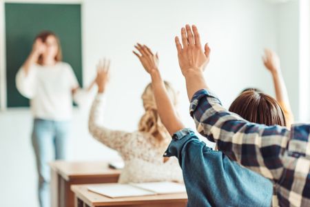 In the foreground, people sit at desks with their hands raised. In front of them a woman in a white sweater stands against a green chalkboard.
