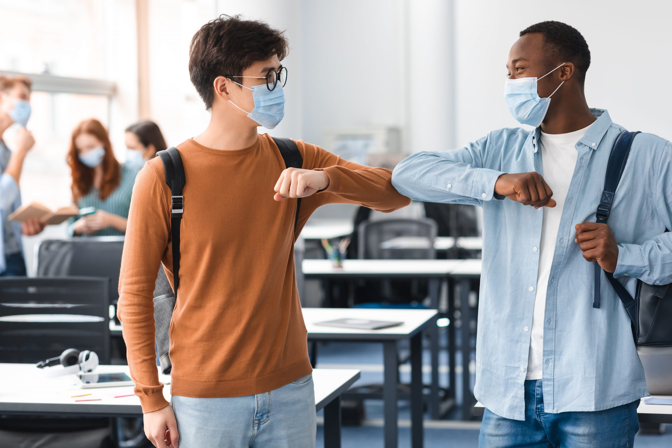 two students standing six feet apart bumping elbows to say hello. Both wearing face masks as they leave their classroom. 