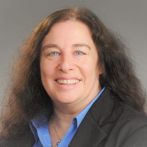 formal headshot of a women with long brown hair wearing a black suit with a blue shirt against a gray background