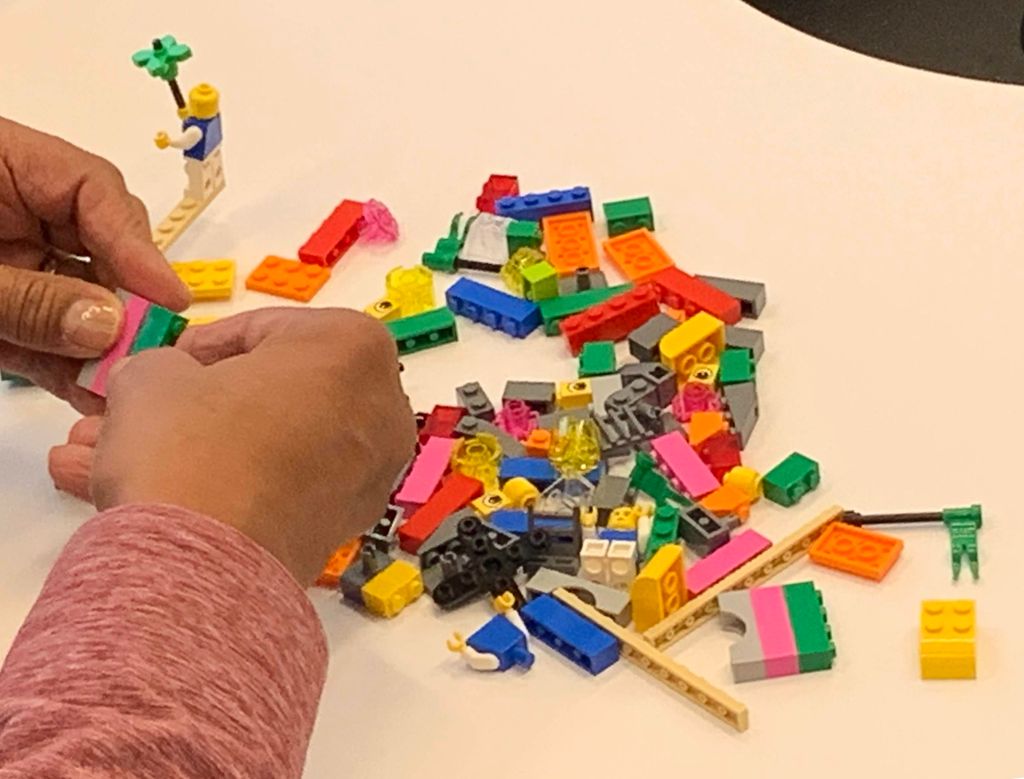 A person's hands fiddle with putting together LEGO bricks that were apparently gleaned from the stack of scattered bricks before them.