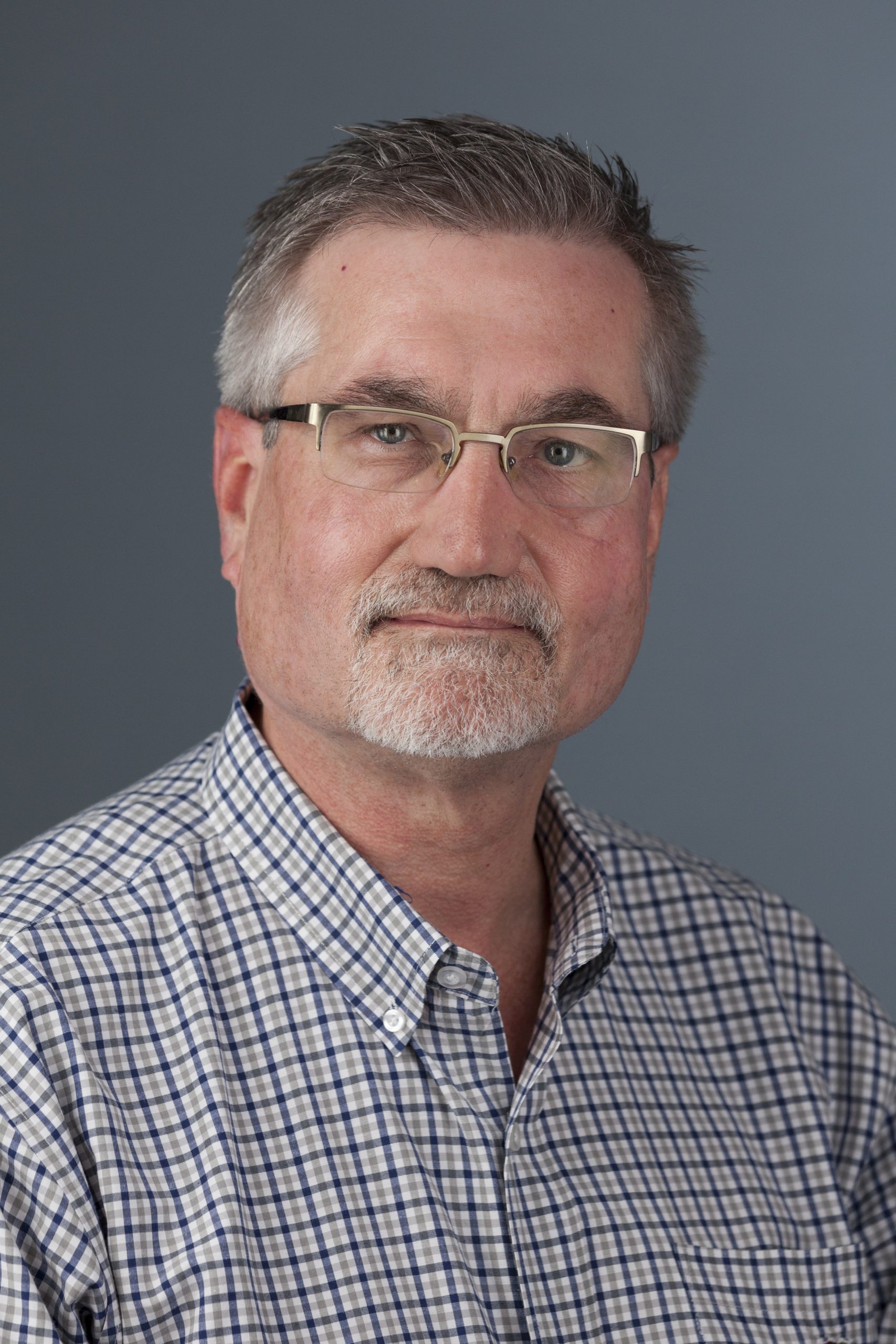 Photo of Michael Wehmeyer wearing glasses and a blue and white checkered shirt in front of a gray background