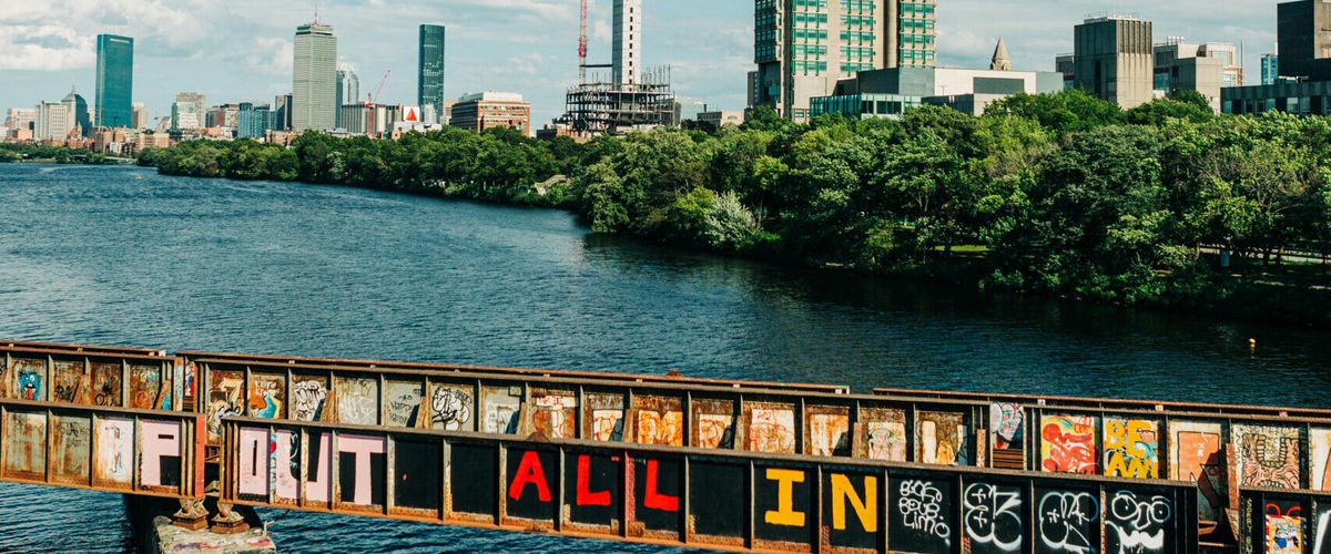 Photo of the Charles River and Boston skyline. Bridge in foreground has spray-painted words "ALL IN"